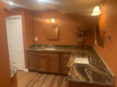 This bathroom showcases Colorado Home Remodeling style and attention to detail, featuring orange walls, two sinks set in marble countertops, wooden cabinets, a vase of dried flowers, and two wall-mounted mirrors with lights.