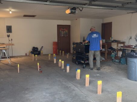 A person stands in a garage and arranges multiple wooden blocks—each topped with yellow paint—in a grid pattern on the floor, possibly preparing for a renovation project such as remodeling a bathroom or basement. Tools and equipment fill the workspace.