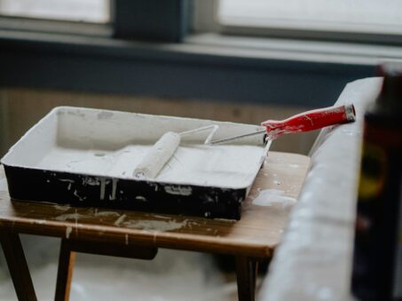 A paint roller with a red handle sits in a tray of white paint on a wooden table by a window, ready for home remodeling.