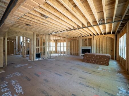 Natural light fills a house under construction, exposing wood framing, unfinished floors, and fireblocking features near a fireplace.