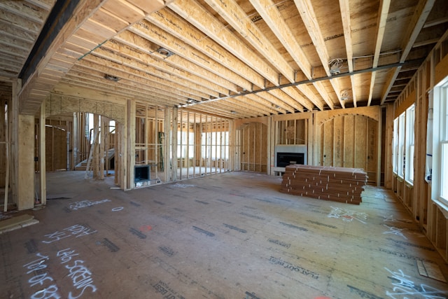 Natural light fills a house under construction, exposing wood framing, unfinished floors, and fireblocking features near a fireplace.