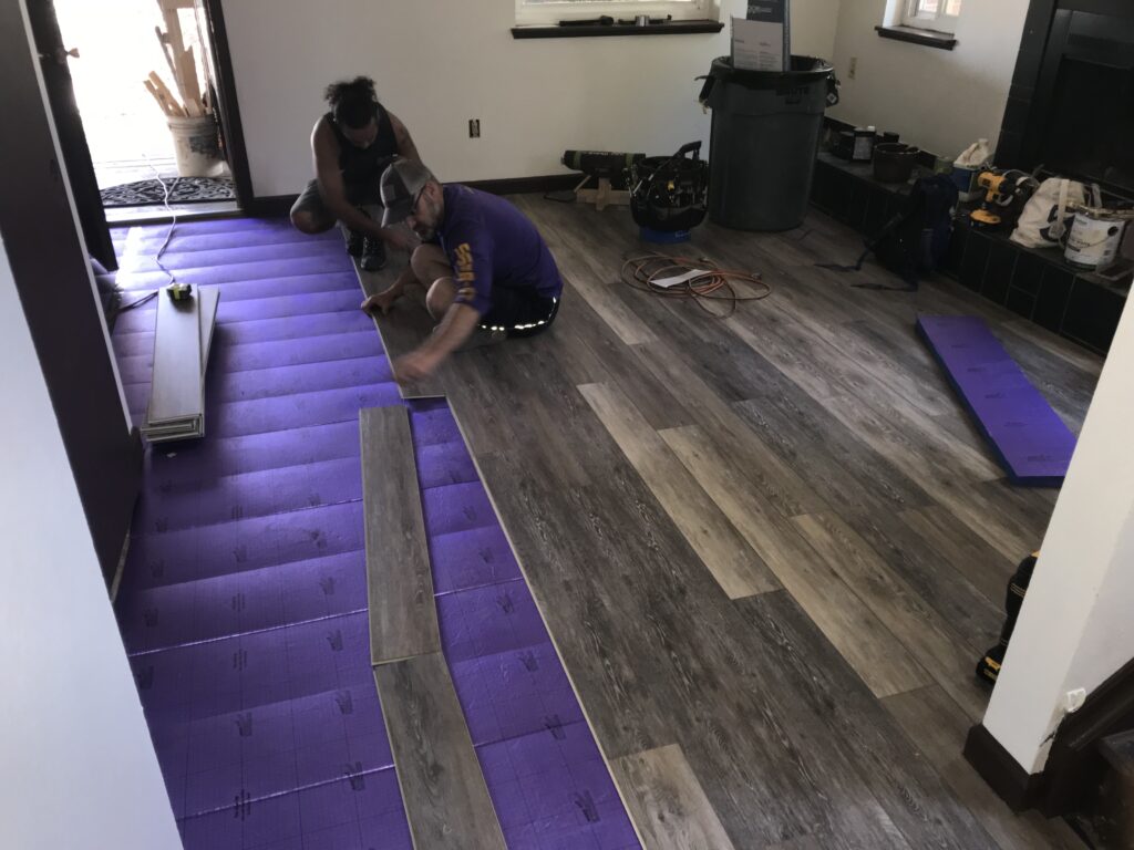 Two people install premium LVP flooring over a purple underlayment in a residential room. Nearby, tools and materials are neatly arranged.