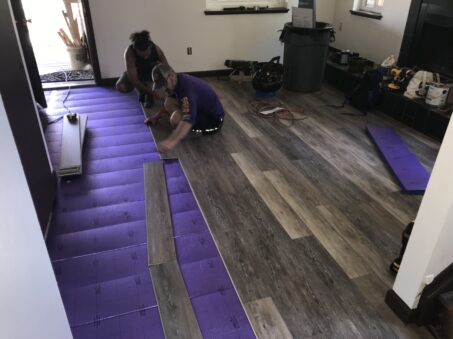 Two people install premium LVP flooring over a purple underlayment in a residential room. Nearby, tools and materials are neatly arranged.