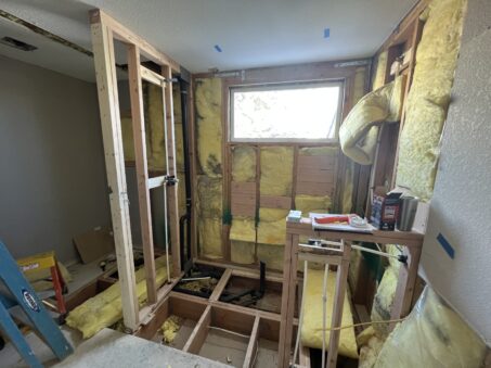 Workers renovate a bathroom, exposing wall studs, insulation, and plumbing pipes while construction materials cover the floor.
